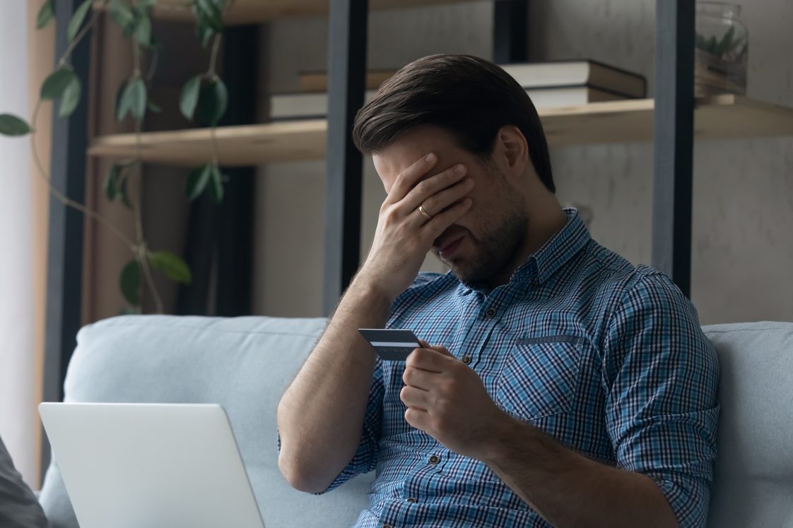 upset man with face in hands looking at his laptop with credit card in hand